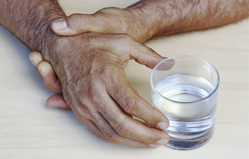 En persons højre hånd, der rækker ud efter et glas vand, mens den venstre hånd holder højre hånd stille, så den ikke ryster.